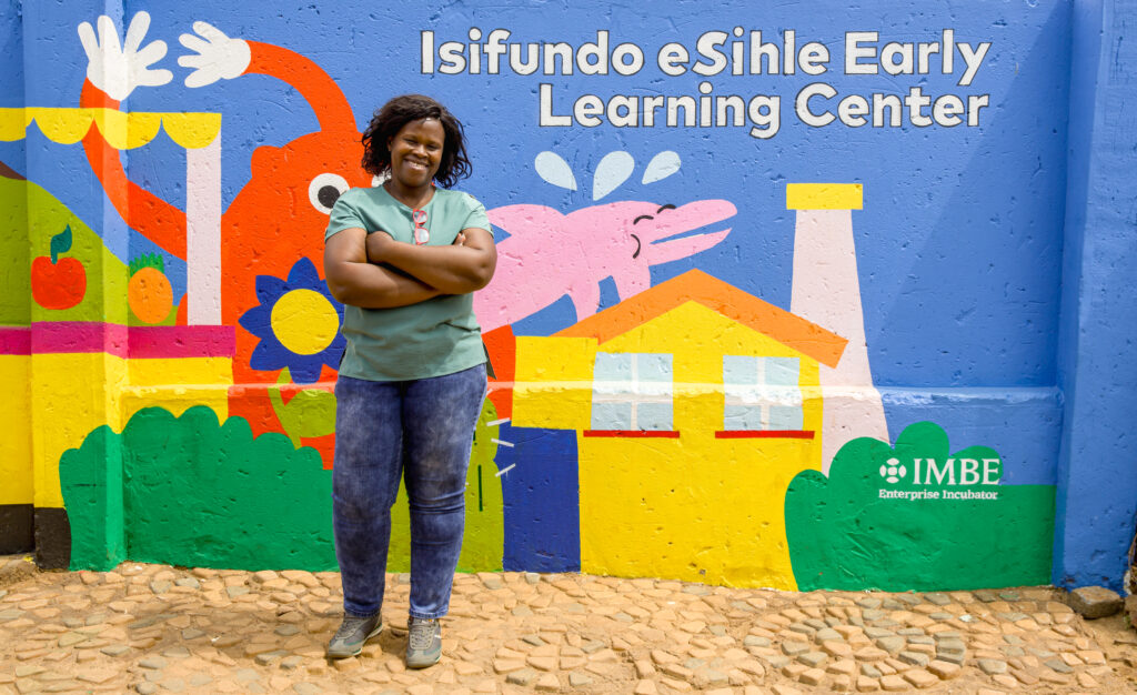 woman standing in front of a wall with the writing "isifundo eShila early learning center"